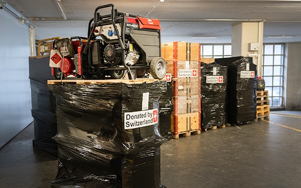 In un magazzino, una scatola con l'immagine di una stufa mobile su un pallet è in primo piano. Sullo sfondo, altri pallet su cui sono posizionati i generatori di corrente imballati.