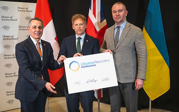 Bundesrat Cassis, der britische Minister Grant Shapps und der Staatssekretär des ukrainischen Aussenministeriums, Oleksandr Bankov, stehen nebeneinander und halten gemeinsam ein Dokument.