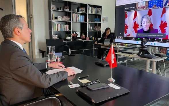 Federal Councillor Cassis sits at a table and looks at a large screen showing a woman giving a speech.