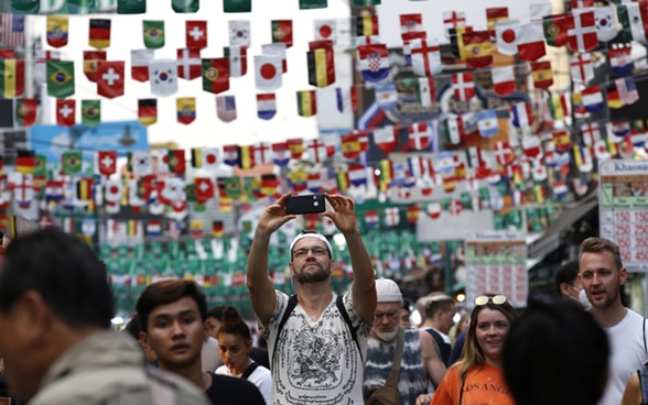 A tourist takes a selfie on a crowded street with flags hanging above.