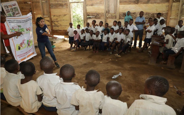 A woman is teaching a group of children about the dangers of an explosive device lying on the ground in the middle of the group.