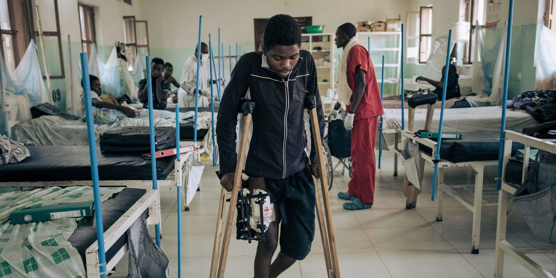 A young African man with a gunshot wound to the leg walks with difficulty on crutches in a hospital.