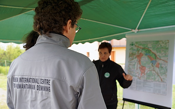 Un homme de dos porte une veste avec la mention GICHD et regarde une femme qui donne des explications sur une carte.