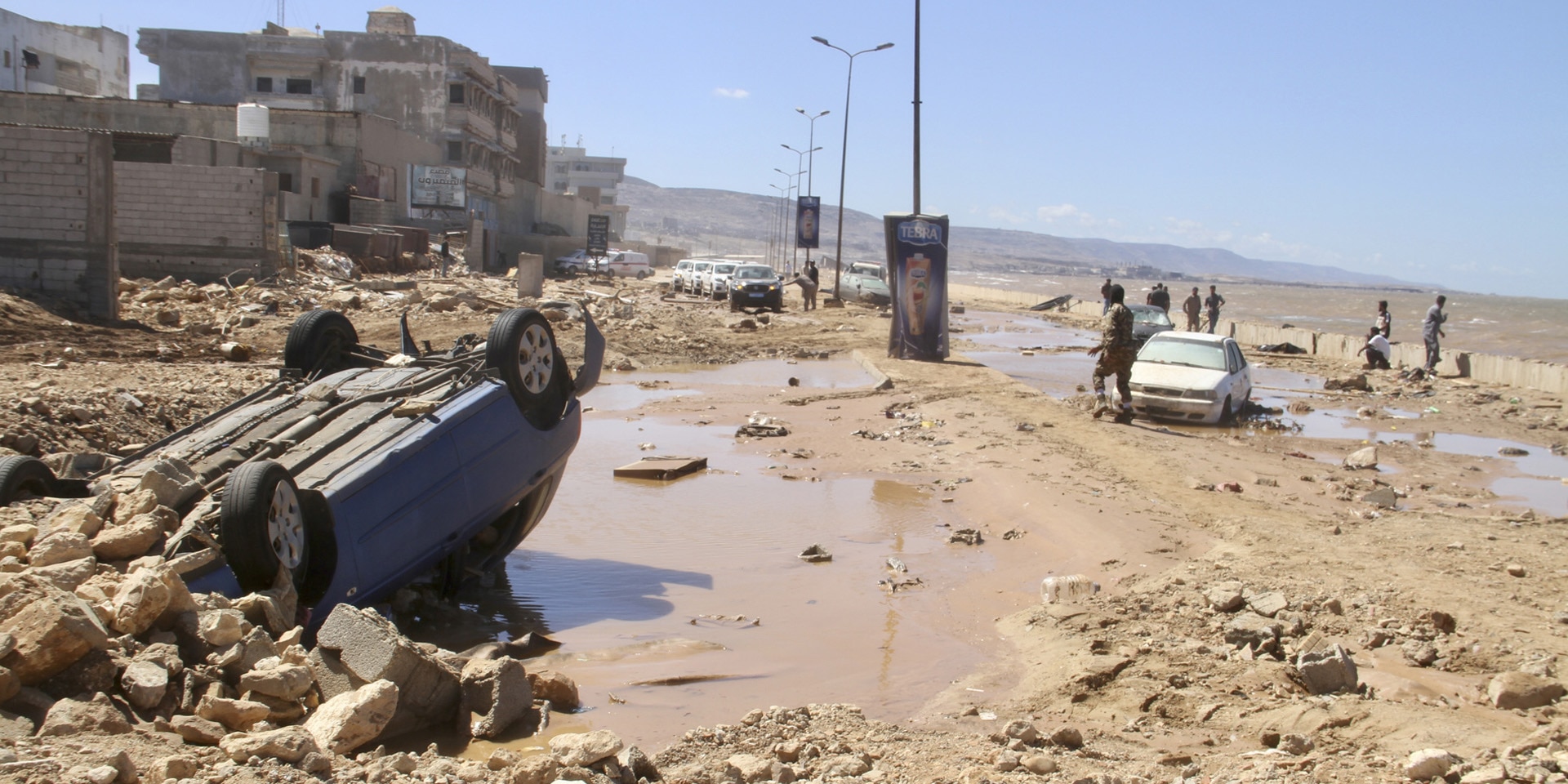 A car lies on its roof in mud and wall remains. People search for victims of the flood in the background.