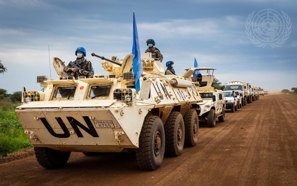 A convoy of white armoured UN vehicles drives along a dusty road.