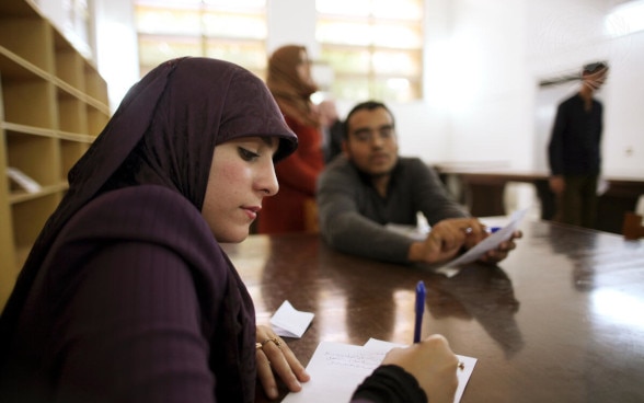 A young Libyan woman wearing a purple headscarf sits at a wooden table taking notes.