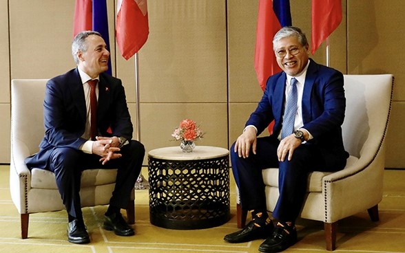 Ignazio Cassis and Enrique Manalo are sitting on two chairs. Behind them the flags of their respective countries.