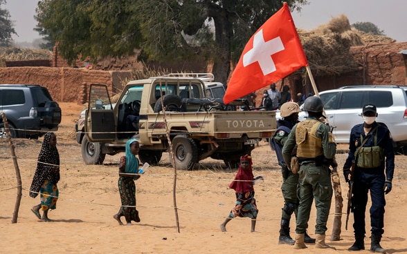 Einige Soldaten stehen neben einem Auto. Eine Person hält eine Schweizer Flagge hoch.