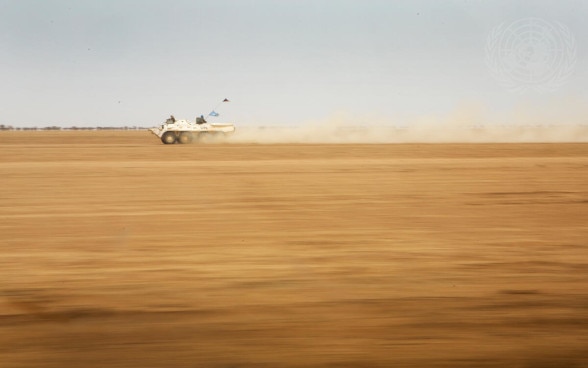 Un véhicule blindé blanc d'une mission de paix de l'ONU roule à grande vitesse dans un paysage désertique.