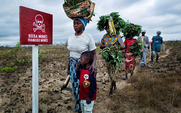 Un gruppo di donne che rientra dal lavoro nei campi passa davanti a un cartello su cui c’è scritto «Pericolo: Mine» ai margini di un terreno agricolo.