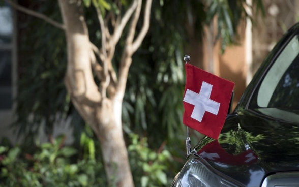 Une voiture arborant un drapeau suisse symbolise la fonction de «facteur» que la Suisse assume en tant que puissance protectrice.