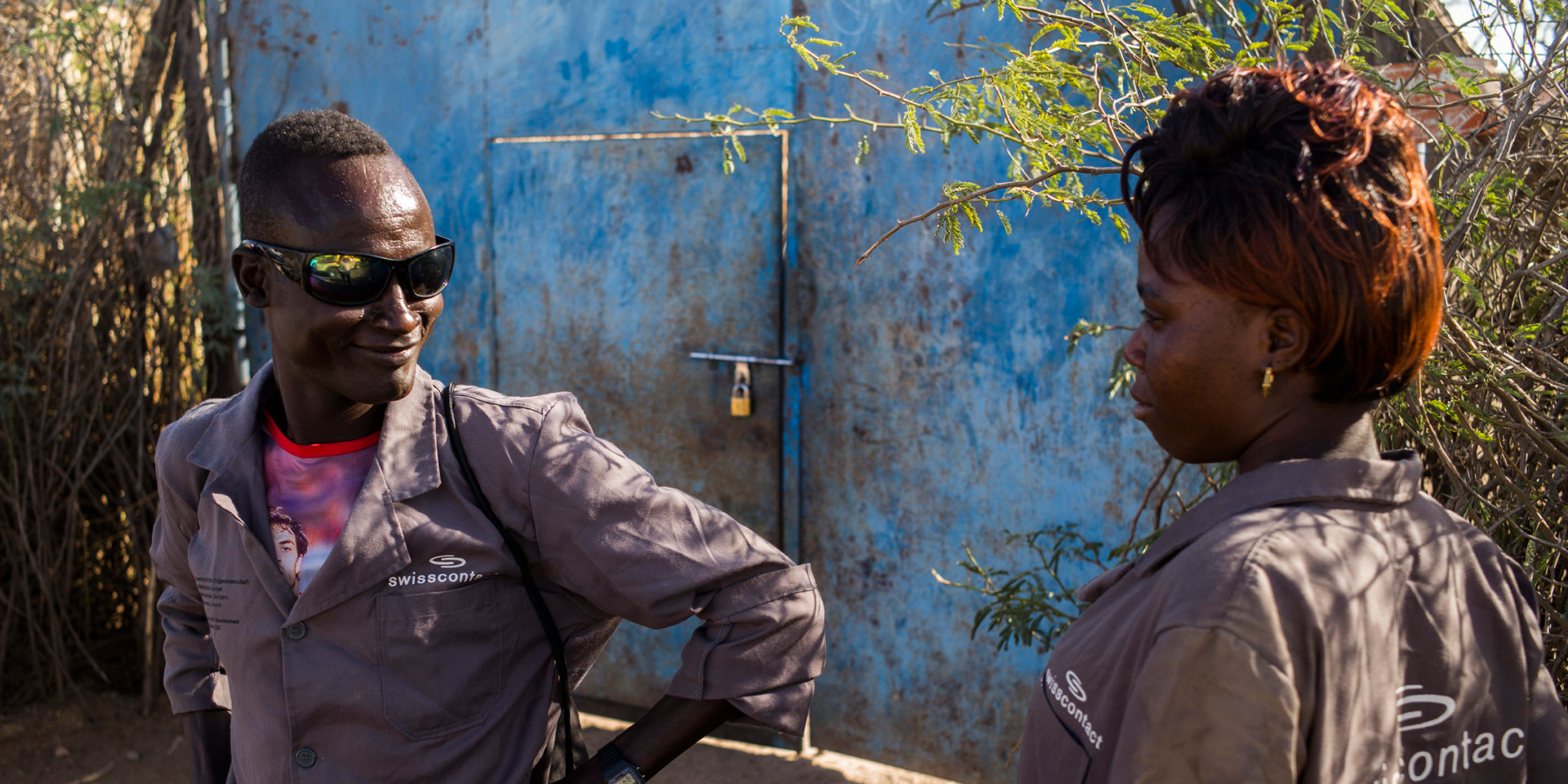 A dark-skinned man wearing sunglasses stands next to a woman in front of a blue door.