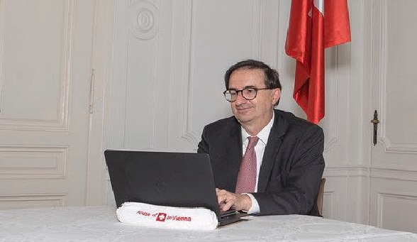 L'ambasciatore Wolfgang Amadeus Brülhart è seduto nel suo ufficio di casa e lavora al portatile. Sullo sfondo è possibile vedere una bandiera svizzera.