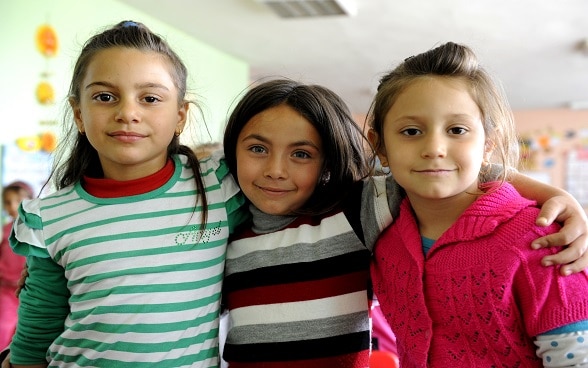 Trois jeunes filles se tiennent par les épaules et sourient au photographe.